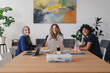 © Studio Marmellata - Three diverse young adults, two women and one man, sit at a table with laptops and notebooks, smiling at the camera. They are engaged in a collaborative work or study session.