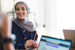 © Studio Marmellata - A smiling young woman in a hijab sits at a desk, holding a pencil. She is attentively participating in a discussion or meeting, with laptops nearby. Her expression is positive and engaged.