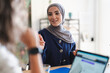 © Studio Marmellata - A smiling young Muslim woman in a hijab engages in a conversation during a meeting. She is seated at a table with a laptop and a blue folder, actively participating.