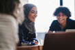 © Studio Marmellata - Diverse young adults, including a woman in a hijab, smile while collaborating at a table. A man looks at a laptop. They share a positive, engaging moment.