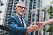 © deagreez - Mature businessman confidently shaking hands outdoors in urban setting wearing formal suit