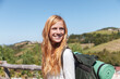 © Marino Bocelli - Young woman smiling, carrying a backpack and sleeping mat, looking away with hills in background