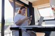 © Shi  - Young woman sitting at desk with laptop, using smartphone for communication and multitasking. Concept of remote work, online study, e-banking, digital lifestyle, mobile technology and productivity