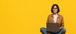 © Prostock-studio - Happy millennial woman working online, sitting on chair and using laptop on yellow studio background, full length. Excited lady surfing internet on portable pc