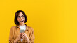 © Prostock-studio - Dreamy young woman using smartphone and looking aside, standing over yellow studio background. Happy lady surfing web, sending sms, chatting to friend, posting in social media