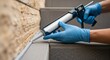 © Margarita Ratatosk - Person in blue gloves using a caulk gun to apply sealant to a building exterior. Home repair and maintenance for weatherproofing concept.