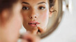 © dominic - Reflective mirror showing woman applying lip gloss, showcasing beauty routine and makeup application in softly lit indoor setting with natural features.