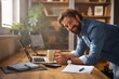 © Stockphotodirectors - A joyful man leans on his desk in a home office, holding a cup of coffee and working intently on his laptop. Natural light fills the space, enhancing the welcoming atmosphere.