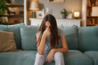© Stockphotodirectors - A teenage girl sits on a soft couch in a warmly lit living room, her head down and hand covering her face, visibly expressing frustration and concern over her current challenges.