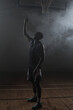 © wavebreak3 - Lone athlete standing under basketball hoop on misty wooden court floor, casting subtle shadows