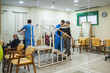 © Koldo_Studio - Healthcare worker helping elderly man exercising on rehabilitation stairs, while other seniors perform physical therapy in a nursing home