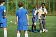 © Mediaphotos - Black man coaching group of teenage boys during soccer training session on outdoor field, demonstrating technique with soccer ball while giving instructions