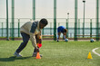 © Mediaphotos - Black man arranging orange cones on outdoor soccer field while teenager in blue sportswear stretching in background, soccer balls scattered on artificial turf