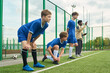 © Mediaphotos - Three teenage boys preparing for soccer training on outdoor field, standing and crouching near sideline while Black man coach giving instructions in background
