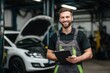 © id512 - Smiling auto mechanic holding digital tablet in repair shop. Male technician standing in front of car with open hood at garage service center.