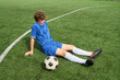 © Mediaphotos - Teenage boy sitting on soccer field resting with soccer ball in front, wearing sports uniform and cleats, looking down with hands on grass, taking break after playing