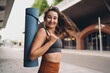 © BullRun - Happy woman smiling with yoga mat over shoulder, hair blowing in wind. Conceptual stock photo symbolizing wellness, mindfulness, empowerment, and connection in digital culture.
