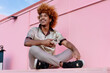 © BullRun - Relaxed African American man with curly ginger hair sitting outdoors holding smartphone, glancing sideways with smile, representing positive attitude toward mobile technology and connectivity.