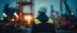 © Ol - The construction worker overlooking an illuminated industrial site with cranes at dusk