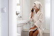 © sutulastock - Woman in bathrobe and towel turban talking on smartphone while wearing hydrating sheet mask in bathroom
