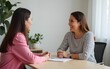 © Donna - Two women sit at light brown table. One woman wears pink top, striped top. Appear to in conversation counseling session. Pro setting suggests possible roles as teacher, therapist counselor. Scene