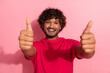 © deagreez - Cheerful young man with curly hair in a red t-shirt smiles and gives a thumbs-up gesture against a bright pink backdrop.