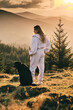 © KatyaPulka - A woman in a sweatsuit standing with her black dog in a mountain landscape at sunset. Golden hour light illuminates the beautiful view. Peaceful, serene moment.