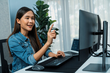 Young Asian girl of IT developer pointing online information on pc with coding program data application on website project, wearing jeans shirt. surrounded by safety programing two screens. Stratagem.