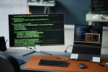Closeup showing computer monitor and laptop displaying programming code on screens, modern workspace setup for software development, no people visible, focus on technology
