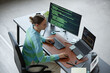 © Seventyfour - Caucasian young adult woman working at desk using desktop computer and laptop with programming code on screens, holding mouse and typing on keyboard, surrounded by office supplies