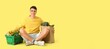 © Pixel-Shot - Young man with full shopping basket and paper bag sitting on yellow background with space for text