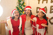 © GAJENDRRA BHATI  - Young indian female friends wearing santa hats celebrating Christmas together in front of a decorated tree at home.