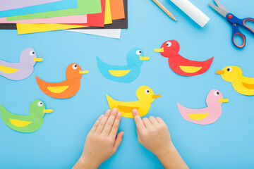 Wall Mural - Little child boy hands creating and gluing duck shapes. Scissors, glue stick, pencil and colorful application paper on blue table background. Pastel color. Closeup. Point of view shot. Top down view.