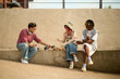 © DragonImages - Group of three young people, including Caucasian man, Caucasian woman, and Black woman, sitting outdoors on concrete ledge eating snacks and drinking from bottles together