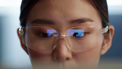 Extreme close up of a focused Asian womans eyes wearing glasses with a computer screen reflecting data code and analytics representing technology research and innovation.