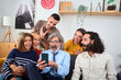 © CarlosBarquero - Caucasian people of different generations sitting on sofa using and looking at cell with amazed faces in living room. Happy family gathered surprised watching videos on the mobile phone at home