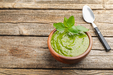 Wall Mural - Tasty green pesto in bowl and basil leaves on wooden table, space for text