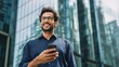 © Yuliia Malovana - Portrait of smiling businessman outside holding smartphone while looking up in modern city environment, dressed in blue shirt. Male employee standing outdoors with phone and glass buildings behind.