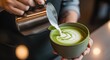 © Karinka - A barista pours steamed milk from a steel pitcher to create latte art on a green matcha tea in a ceramic cup.