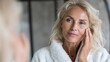 © Johannes - gorgeous mid age older adult 50 years old blonde woman wears bathrobe in bathroom applying nourishing antiage face skin care cream treatment looking at mirror doing daily morning beauty routine no lo