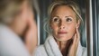© Johannes - gorgeous mid age older adult 50 years old blonde woman wears bathrobe in bathroom applying nourishing antiage face skin care cream treatment looking at mirror doing daily morning beauty routine no lo