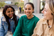 © Xavier Lorenzo - Group of three diverse student friends is sitting on a bench at the university campus, talking and laughing together. Latin American girl smile at camera. Friendship and youth community concept
