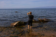 © Светлана Высокос - A white woman with blonde hair stands on a rocky shore, looking at calm water and distant mountains.