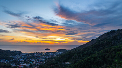 Wall Mural - A breathtaking sunset paints the sky in vivid hues of orange and blue over a coastal town, with lush green hills framing the scene. A distant hilltop structure adds a serene tranquil landscape.