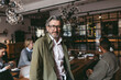 © Maskot - Portrait of confident senior male business professional wearing eyeglasses standing near table in office