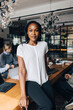 © Maskot - Portrait of confident young female business professional sitting at edge of table in office