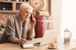 © Prostock-studio - Senior Businesswoman Using Laptop, Talking On Cellphone And Taking Notes While Working From Home Sitting In Kitchen
