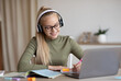 © Prostock-studio - Smiling teen girl in wireless headset having online lesson while staying home during coronavirus pandemic, using laptop and taking notes in her exercise book, copy space. Home schooling concept