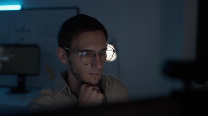 Male software developer wearing glasses coding in dark office at night. Man looking screen desktop remote work overtime in blue light at home.