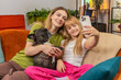© Andrii Iemelianenko - Mother and daughter with French bulldog at home use smartphone selfie filter mask joke laugh smile. Happy woman and girl with dog on sofa make faces take photo share moment family joy vibe together